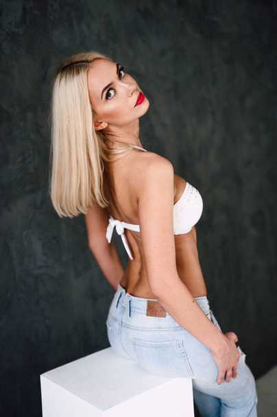 glamouröse junge sexy blonde Frau trägt weißen BH und Jeans mit roten Lippen auf dunklem Hintergrund - Foto, Bild
