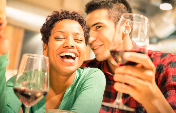 Happy multiracial couple of lover drinking red wine at fashion restaurant - Handsome man whispers soft kisses in beautiful woman ear - Relationship concept with boyfriend and girlfriend on warm filter - Photo, image