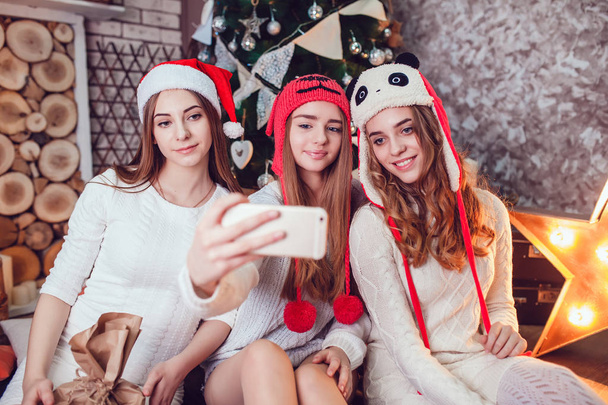 Beautiful girls in funny hats sitting on the floor near a Christmas tree and make selfie. New Year. - Valokuva, kuva