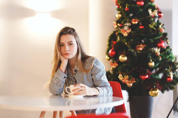 Young beautiful woman drinks hot coffee in cafe with Christmas tree decorations - Foto, Imagen