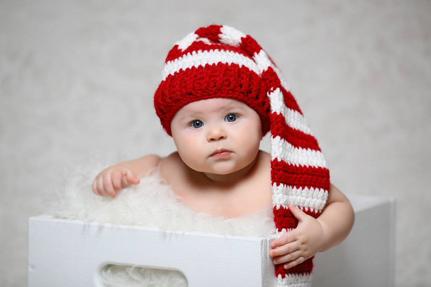Jouluvauva istuu valkoisessa puulaatikossa punainen ja valkoinen raidallinen pitkähäntäinen tonttuhattu päässä.
 - Valokuva, kuva