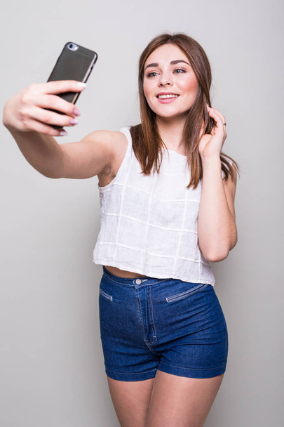 jong meisje maakt selfie met smartphone geïsoleerd op een grijze achtergrond - Foto, afbeelding