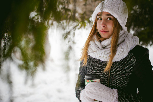 Portrait of cute woman in white scarf and hat knitted coat on outdoors background of snow and blurred fir branches in winter. - Foto, Bild
