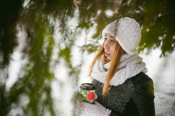 Portrait of cute woman in white scarf and hat knitted coat on outdoors background of snow and blurred fir branches in winter. girl holds a coffee cup in hand, christmas takeaway coffee - Fotoğraf, Görsel
