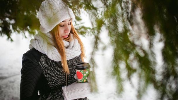 Retrato de mujer linda en bufanda blanca y sombrero abrigo de punto en el fondo al aire libre de la nieve y ramas de abeto borrosas en invierno
. - Foto, Imagen