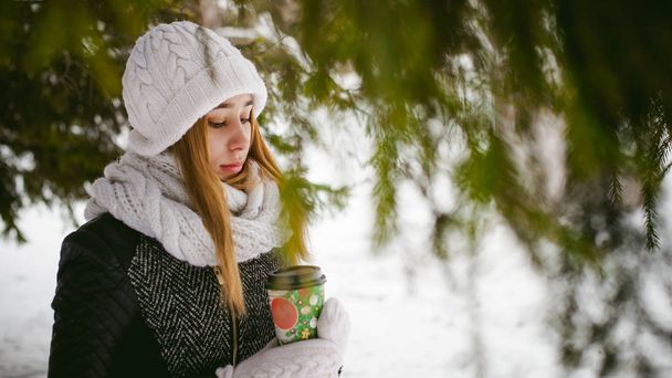 Portrait of cute woman in white scarf and hat knitted coat on outdoors background of snow and blurred fir branches in winter - Photo, image