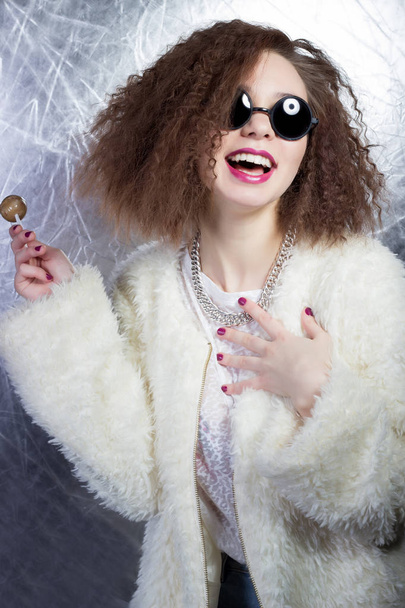 funny mischievous cheerful girl smiling with curly hair in a white blouse and jeans in the sun round glasses with candy in studio - Zdjęcie, obraz