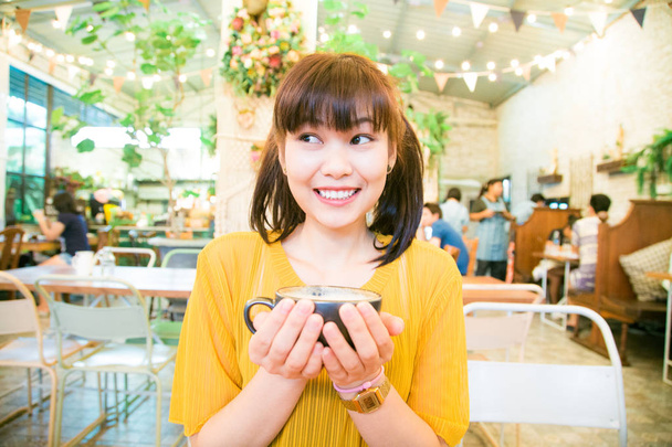 S úsměvem asijské ženy posezení v umělecké kavárně drží latte černý hrnek - Fotografie, Obrázek