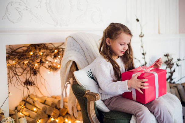 Gelukkig kind meisje zittend op fauteuil bedekt met een deken tegen ingerichte kerst open haard - Foto, afbeelding