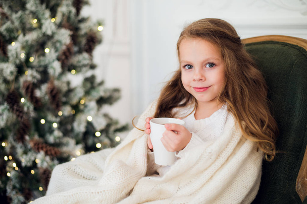 Little girl wearing pajama sitting on armchair wrapped in a blanket with holiday bedding by Christmas tree and fireplace  drinking milk at home - Photo, image
