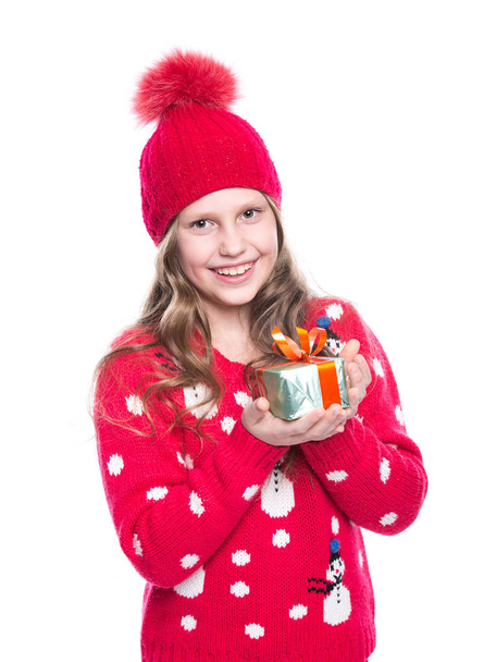 Charming smiling little girl with curly hairstyle wearing red knitted sweater and hat holding christmas gift isolated on white background. Winter clothes and christmas concept. - Photo, Image