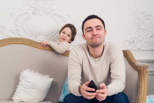 zmęczony zdenerwowany ojciec siedzi na kanapie przy użyciu telefonu i syna, który chce grać z nim - Zdjęcie, obraz