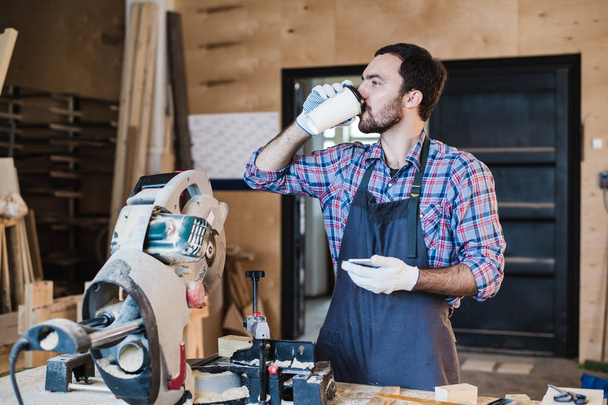 Kávový hrnek za dobrou práci. Promyšlené mladých mužů carpenter drží pohár stoje u dřevěného stolu s rozmanitou pracovní nástroje, kterým na něm - Fotografie, Obrázek