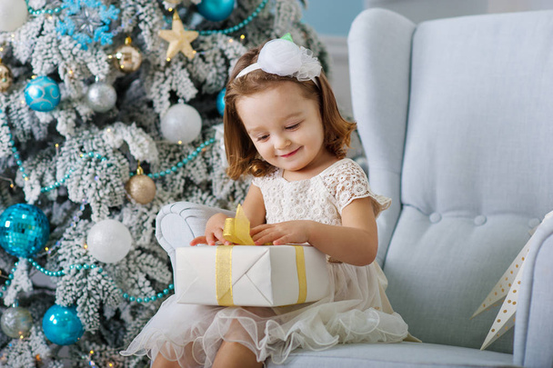 cute little girl in bklom dress sitting in a chair and opens box with present for background Christmas tree blue ornaments - Zdjęcie, obraz