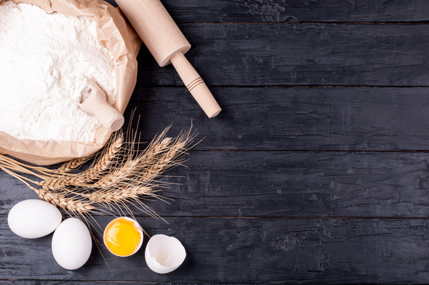 Ingredients for baking. Flour in paper bag, wheat eggs and kitchen utensils - rolling pin and whisk on dark wooden background. Cooking cookies. Top view - Photo, Image