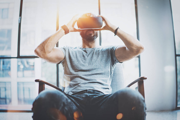 Attractive bearded man enjoyingvirtual reality glasses in modern interior design coworking studio.Home play concept.Smartphone use with VR goggles headset. Front view,flare effect,blurred background. - 写真・画像