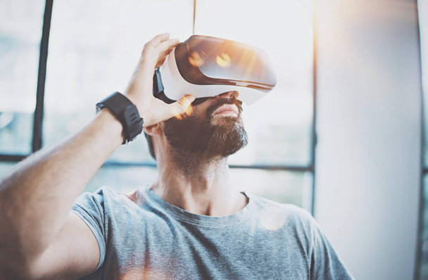 Attractive bearded man enjoyingvirtual reality glasses in modern interior design coworking studio.Home play concept.Smartphone use with VR goggles headset.Flare and sunny effect,blurred background. - Φωτογραφία, εικόνα