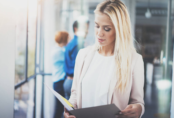 Portrait of young pretty blonde businesswoman in modern coworking studio with business team on the background.Beautiful woman near the window holding papers in hands.Horizontal,blurred. - Foto, Imagen