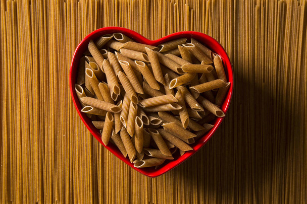 Wholemeal Pasta. Integral Penne into a heart bowl over a wooden  - Fotó, kép
