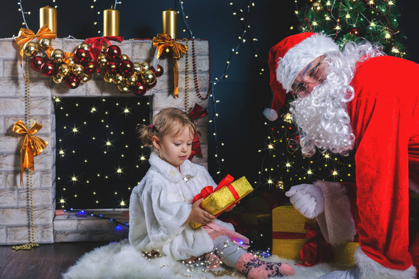 Santa and girl playing near fireplace, decorated Christmas tree - Foto, afbeelding