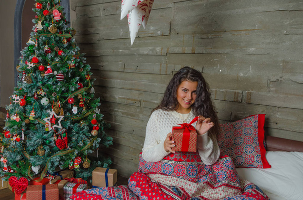 the girl in the white sweater in a warm bed unwrapping Christmas gifts. - Foto, imagen