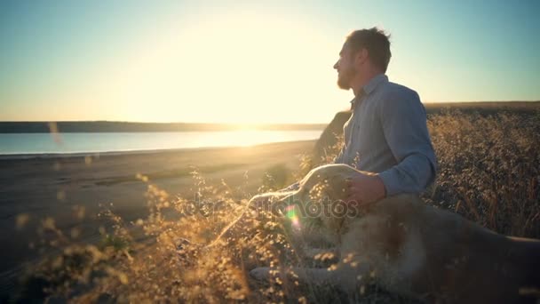 Χαμογελαστός άνθρωπος και σκύλος κάθεται στην άκρη του γκρεμού στο ηλιοβασίλεμα - Πλάνα, βίντεο