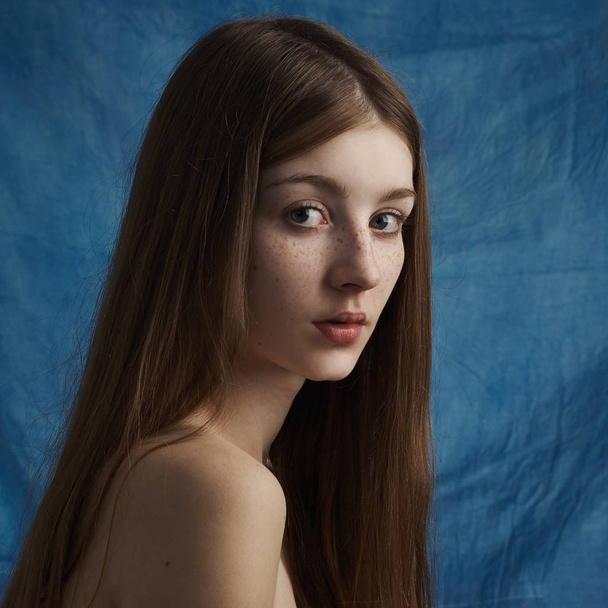 Beauty theme: portrait of a beautiful young girl with freckles on her face and wearing a black dress on a blue background in studio - Zdjęcie, obraz