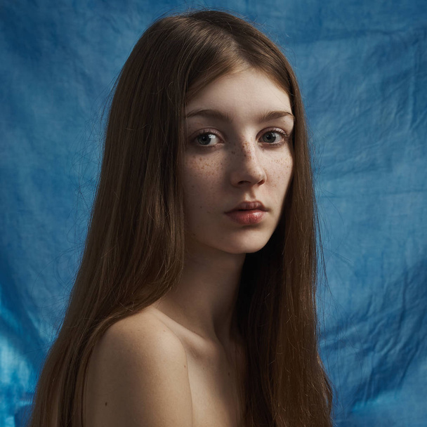 Beauty theme: portrait of a beautiful young girl with freckles on her face and wearing a black dress on a blue background in studio - Zdjęcie, obraz