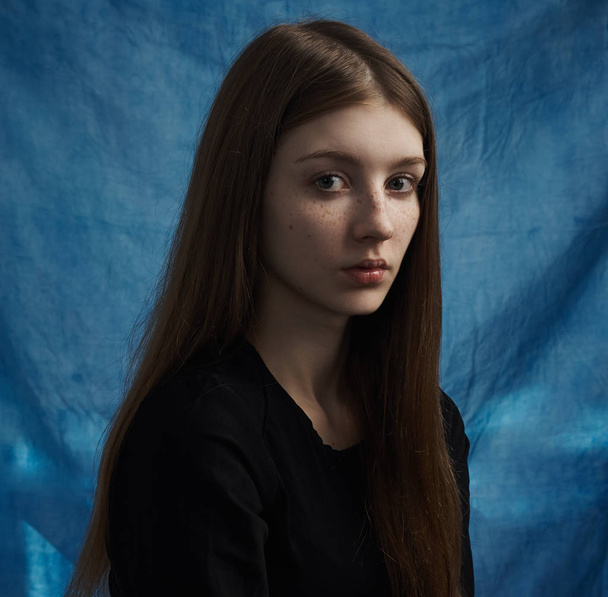 Beauty theme: portrait of a beautiful young girl with freckles on her face and wearing a black dress on a blue background in studio - Foto, Imagem