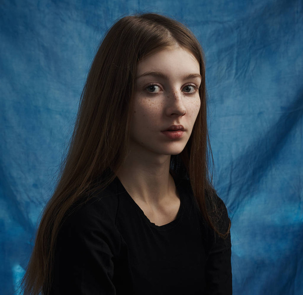 Beauty theme: portrait of a beautiful young girl with freckles on her face and wearing a black dress on a blue background in studio - Foto, imagen