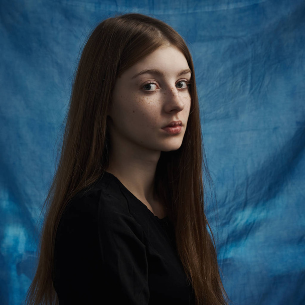 Beauty theme: portrait of a beautiful young girl with freckles on her face and wearing a black dress on a blue background in studio - Foto, Imagem