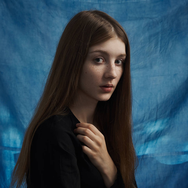 Beauty theme: portrait of a beautiful young girl with freckles on her face and wearing a black dress on a blue background in studio - Photo, Image