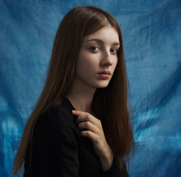 Beauty theme: portrait of a beautiful young girl with freckles on her face and wearing a black dress on a blue background in studio - Fotó, kép