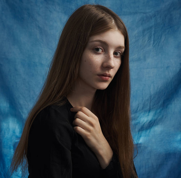Beauty theme: portrait of a beautiful young girl with freckles on her face and wearing a black dress on a blue background in studio - Φωτογραφία, εικόνα