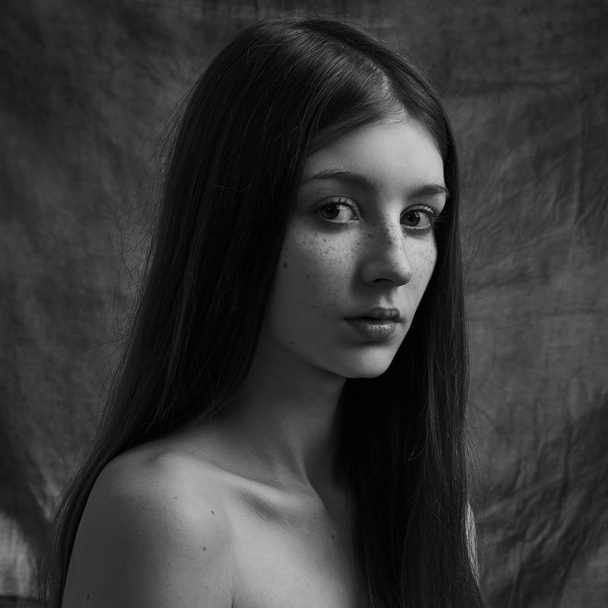 Dramatic black and white portrait of a beautiful lonely girl with freckles isolated on a dark background in studio shot - Zdjęcie, obraz