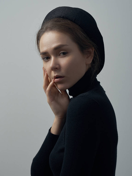 Dramatic portrait of a young beautiful girl with freckles in a black turtleneck and a hat on her head on a white background in studio - Foto, Imagen