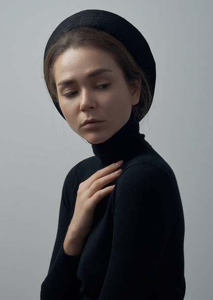 Dramatic portrait of a young beautiful girl with freckles in a black turtleneck and a hat on her head on a white background in studio - Valokuva, kuva