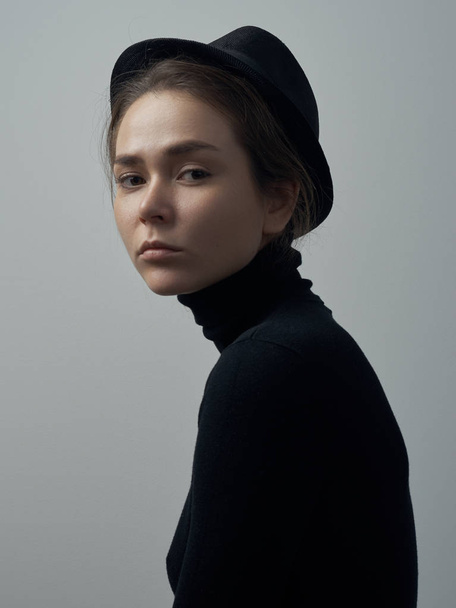 Dramatic portrait of a young beautiful girl with freckles in a black turtleneck and a hat on her head on a white background in studio - Фото, зображення
