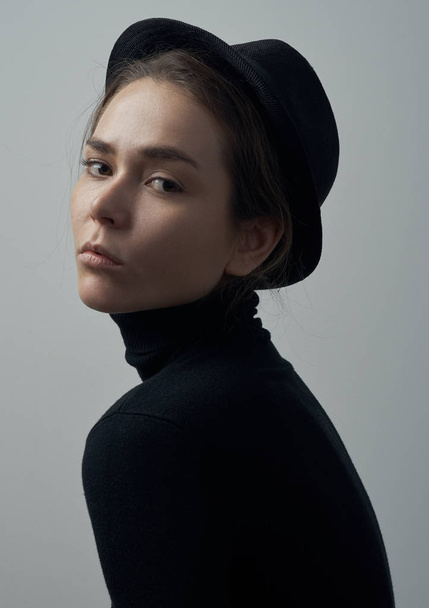 Dramatic portrait of a young beautiful girl with freckles in a black turtleneck and a hat on her head on a white background in studio - Foto, imagen