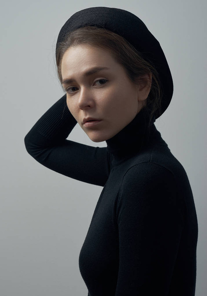 Dramatic portrait of a young beautiful girl with freckles in a black turtleneck and a hat on her head on a white background in studio - Foto, Imagem