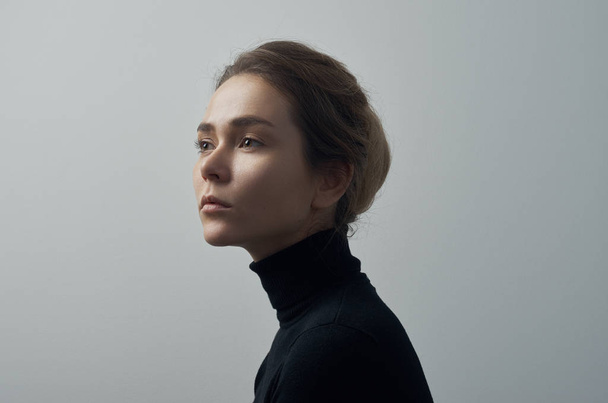 Dramatic portrait of a young beautiful girl with freckles in a black turtleneck on white background in studio - Photo, image