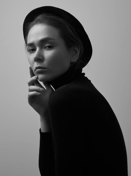 Dramatic black and white portrait of young beautiful girl with freckles in a black turtleneck and a hat on a white background in studio - Фото, изображение