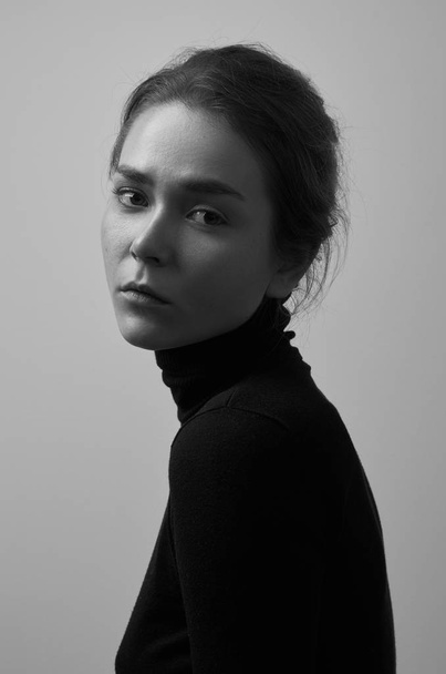 Dramatic black and white portrait of young beautiful girl with freckles in a black turtleneck on white background in studio - Фото, зображення