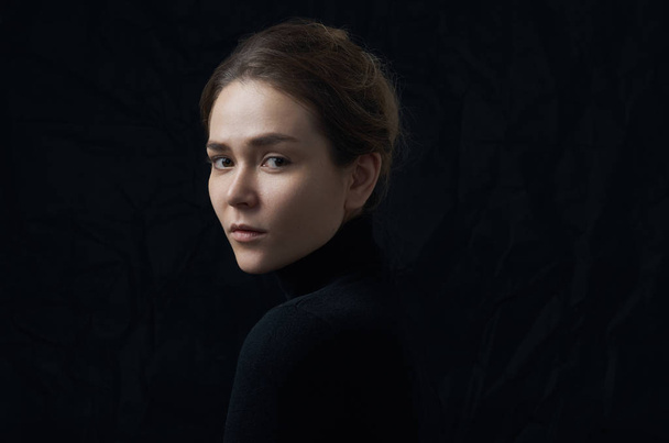 Dramatic portrait of a young beautiful girl with freckles in a black turtleneck on black background in studio - Fotó, kép