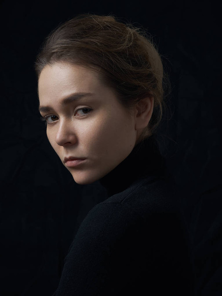 Dramatic portrait of a young beautiful girl with freckles in a black turtleneck on black background in studio - 写真・画像