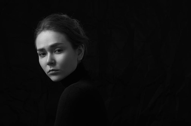 Dramatic black and white portrait of young beautiful girl with freckles in a black turtleneck on black background in studio - Valokuva, kuva