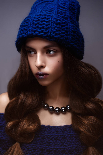 Beautiful girl in winter knitted hat blue color with a necklace around the neck of pearls. Young model with gentle make-up and silver lips. - Φωτογραφία, εικόνα