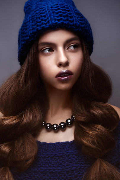 Beautiful girl in winter knitted hat blue color with a necklace around the neck of pearls. Young model with gentle make-up and silver lips. - Foto, Imagen