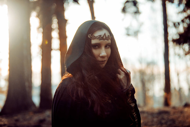 Young beautiful and mysterious woman in woods, in black cloak with hood, image of forest elf or witch - Fotografie, Obrázek
