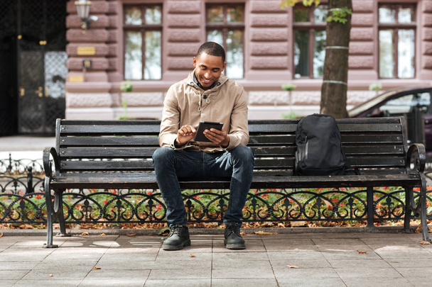 Man with tablet sitting on bench near backpack and typing - Foto, Imagem